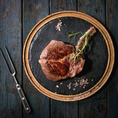 Izgara siyah angus sığır eti tomahawk biftek kemik ile tuz, biber ve biberiye yuvarlak ahşap arduvaz kesme tahtası üzerinde et çatalı koyu ahşap arka plan üzerinde devam eder. Düz yatıyordu. Kare görüntü