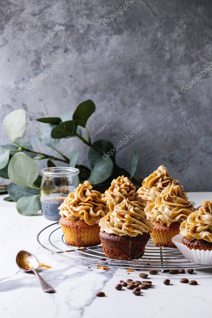 Fresh baked homemade cupcakes with coffee buttercream and caramel standing on cooling rack with eucalyptus branch and coffee beans above over white marble kitchen table.