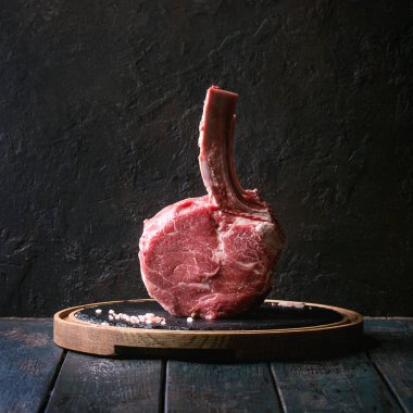 Çiğ çiğ siyah angus sığır eti tomahawk biftek kemik üzerindeki koyu ahşap tahta masa üzerinde tuz ve biber yuvarlak ahşap arduvaz kesme tahtası üzerinde ile görev yaptı. Rustik tarzı. Kare görüntü