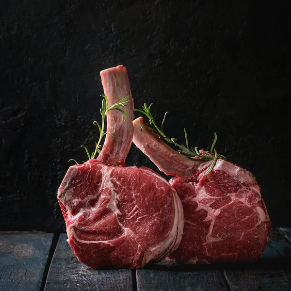 Raw uncooked black angus beef tomahawk steaks on bones served with rosemary over dark wooden plank table. Rustic style. Close up