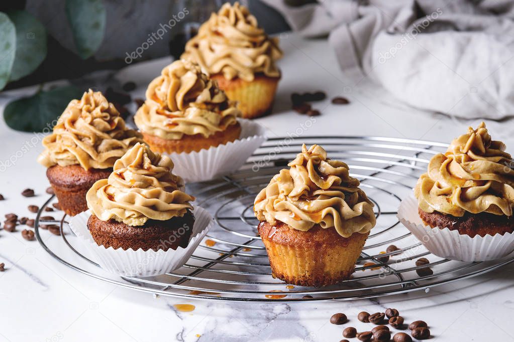 Fresh baked homemade cupcakes with coffee buttercream and caramel standing on cooling rack with eucalyptus branch and coffee beans above over white marble kitchen table.