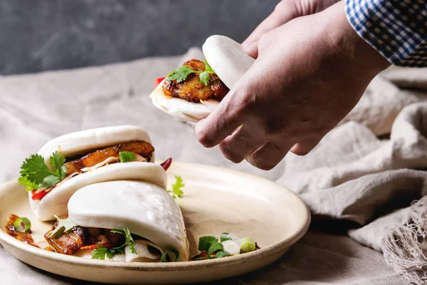 Adamın elleri Asya sandviç buğulama gua bao buns domuz eti, yeşil ve sebzeler ile keten masa örtüsü seramik tabak masaya servis ile tutun. Asya tarzı hızlı yiyecek yemek.