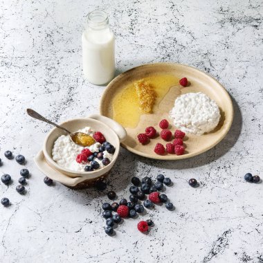 Ev yapımı peynir seramik kase beyaz mermer doku arka plan üzerinde yaban mersini, ahududu, şişe süt ve peteğin ile görev yaptı. Uzay kopyalayın. Kare görüntü