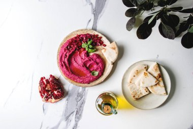 Ev yapımı yayılan kırmızı pancar humus nar taneleri, zeytin yağı, fesleğen ile beyaz mermer arka plan üzerinde pide ekmek ve yeşil Şubesi ile seramik plaka üzerinde görev yaptı. Düz lay, alan. Akdeniz snack.