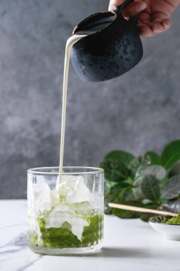 Krema dökerek sürahi dan matcha Yeşil Çay Iced latte veya kokteyl cam, buz küpleriyle matcha toz yeşil dallar tarafından dekore edilmiş beyaz mermer masada. Arka plan gri duvara