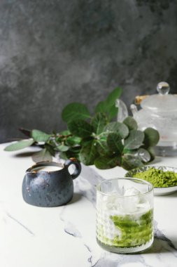 Matcha yeşil latte buzlu çay veya kokteyl cam buz küpleri ile matcha tozu ve süt sürahi beyaz mermer masa, yeşil dallar tarafından dekore edilmiş. Arka plan gri duvara