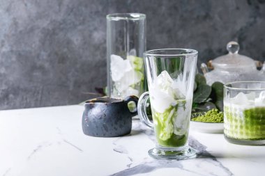 Matcha yeşil latte buzlu çay veya kokteyl buz küpleri ile üç farklı bardaklarda, matcha tozu ve süt sürahi beyaz mermer masa, yeşil dallar tarafından dekore edilmiş. Arka plan gri duvara