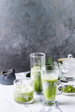 Matcha yeşil latte buzlu çay veya kokteyl buz küpleri ile üç farklı bardaklarda, matcha tozu ve süt sürahi beyaz mermer masa, yeşil dallar tarafından dekore edilmiş. Arka plan gri duvara