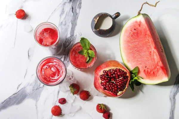 Three different red fruit berry watermelon, strawberry, raspberry, pomegranate cocktails or smoothies in glasses with crushed ice, fresh mint, ingredients above over white marble background. Flat lay