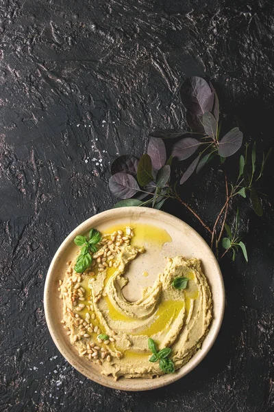 Ev yapımı geleneksel yayılmış humus çam fıstığı, zeytin yağı, fesleğen ile karanlık doku arka plan üzerinde yeşil Şubesi ile seramik plaka üzerinde görev yaptı. Düz lay, alan. Akdeniz snack.