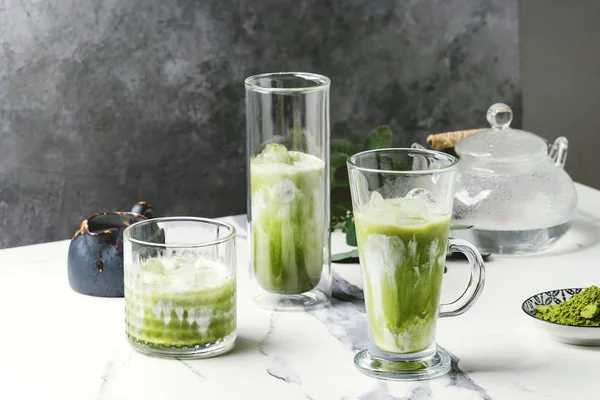 Matcha yeşil latte buzlu çay veya kokteyl buz küpleri ile üç farklı bardaklarda, matcha tozu ve süt sürahi beyaz mermer masa, yeşil dallar tarafından dekore edilmiş. Arka plan gri duvara