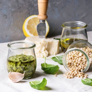 Geleneksel fesleğen pesto sos taze fesleğen, zeytinyağı, parmesan peyniri, sarımsak, çam fıstığı, limon ile beyaz keten masa örtüsü mutfak masasında yukarıdaki malzemelerle cam kavanoza. Uzay kopyalayın. Kare görüntü