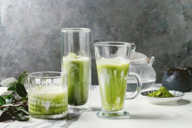 Matcha yeşil latte buzlu çay veya kokteyl buz küpleri ile üç farklı bardaklarda, matcha tozu ve süt sürahi beyaz mermer masa, yeşil dallar tarafından dekore edilmiş. Arka plan gri duvara