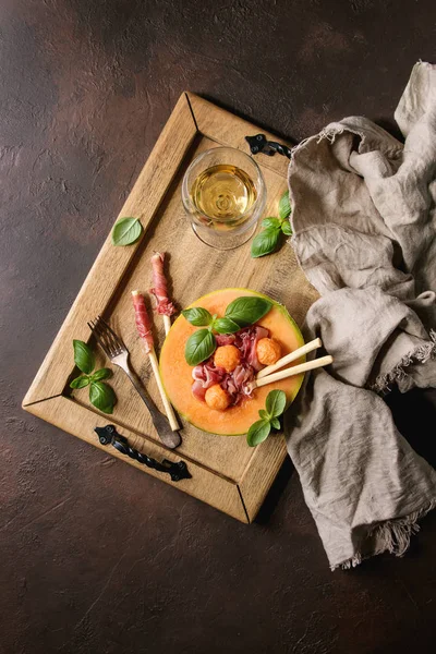 瓜和火腿沙拉的一半甜瓜与新鲜罗勒和 Grissini 面包在木托盘上的黑褐色纹理背景与玻璃白葡萄酒 — 图库照片