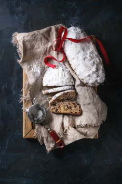 Noel kek dilimlenmiş, geleneksel Alman şenlikli pişirme. Kepekli stollen kuru üzüm ve keten peçete, ahşap tepsi, elek, koyu mavi arka plan üzerinde kırmızı kurdele üzerine pudra şekeri ile. Üstten görünüm, uzay