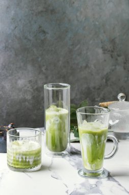 Matcha yeşil latte buzlu çay veya kokteyl buz küpleri ile üç farklı bardaklarda, matcha tozu ve süt sürahi beyaz mermer masa, yeşil dallar tarafından dekore edilmiş. Arka plan gri duvara