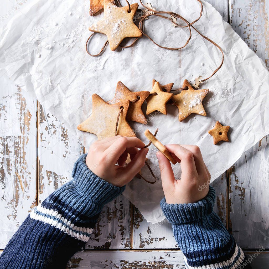 Child hands make garland of homemade shortbread star shape sugar cookies different size on thread on baking paper over white wooden plank table. Christmas handmade gift. Top view, space. Square image