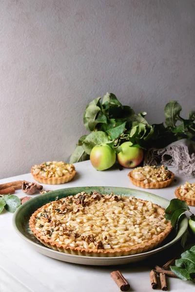 Homemade sweet apple shortbread tart in green ceramic plate and tartlets with cinnamon sticks, walnuts, apples branches above on white marble table. Autumn baking.