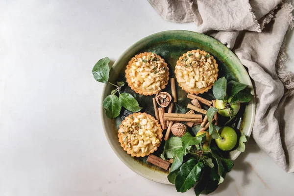 Homemade sweet apple shortbread tartlets in green ceramic plate on cloth with cinnamon sticks, walnuts, apples branches above over white marble background. Autumn baking. Flat lay, space