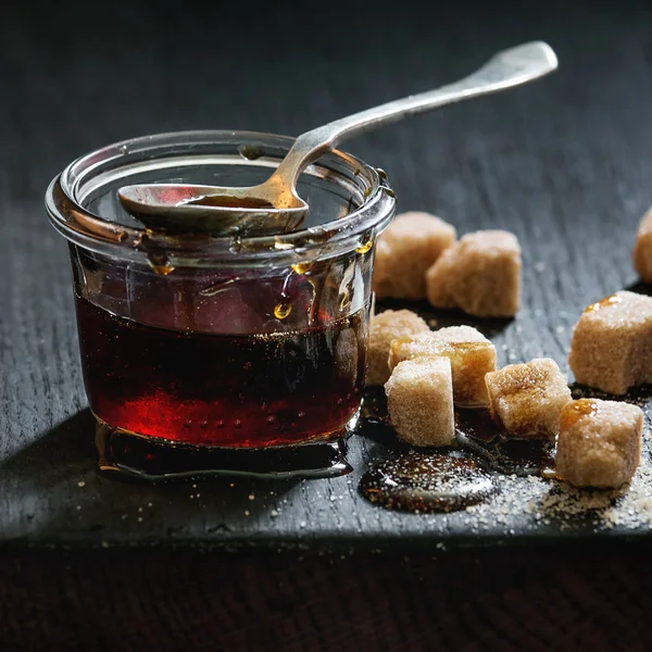 Homemade liquid transparent brown sugar caramel in glass jar standing on black wooden board with spoon and can sugar cubes. Close up. Day light. Square image