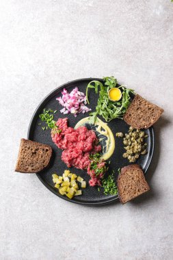 Biftek tartare Bıldırcın yumurta kabuğu, ekmek, turşusu kesim salatalık, kapari, kırmızı soğan, frenk soğanı, roka salatası ile gri arka plan üzerinde siyah seramik tabak içinde görev yaptı. Düz lay, uzay