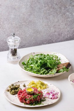 Biftek tartare ile Bıldırcın yumurta kabuğu, çavdar ekmeği, turşusu kesim salatalık, kapari, kırmızı soğan, chives ve roka salatası karabiber değirmeni ile seramik plakalar benekli beyaz mermer masaya servis.