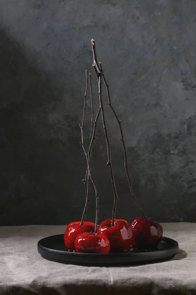 Red caramel apples sweet autumn or Christmas dessert served with branches in black ceramic plate on linen table cloth with grey wall at background. Dark atmospheric mood
