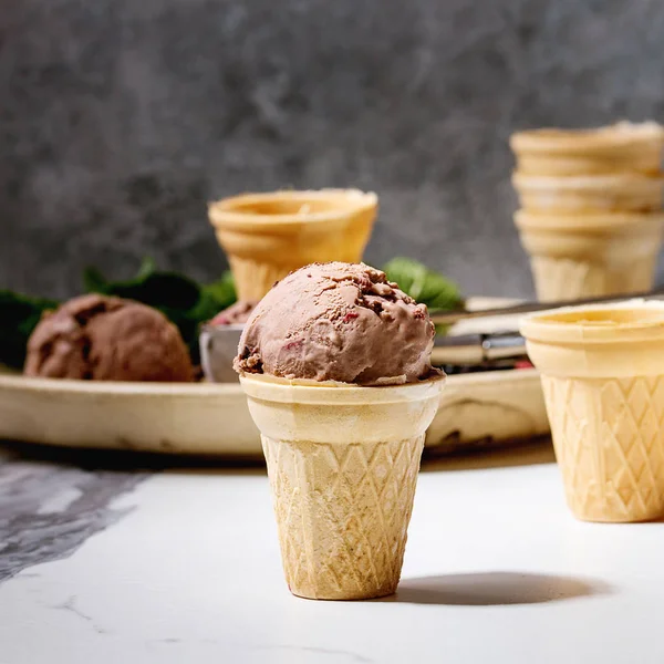 自制的巧克力覆盆子冰淇淋在小华夫饼杯与冷冻浆果 新鲜薄荷 切碎的黑巧克力和金属勺子在陶瓷板在白色大理石厨房的桌子上 正方形图像 — 图库照片