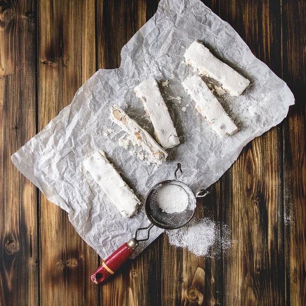 Homemade honey nuts nougat turron sliced on crumpled paper with vintage sieve and sugar powder over dark wooden plank background. Flat lay, space. Square image