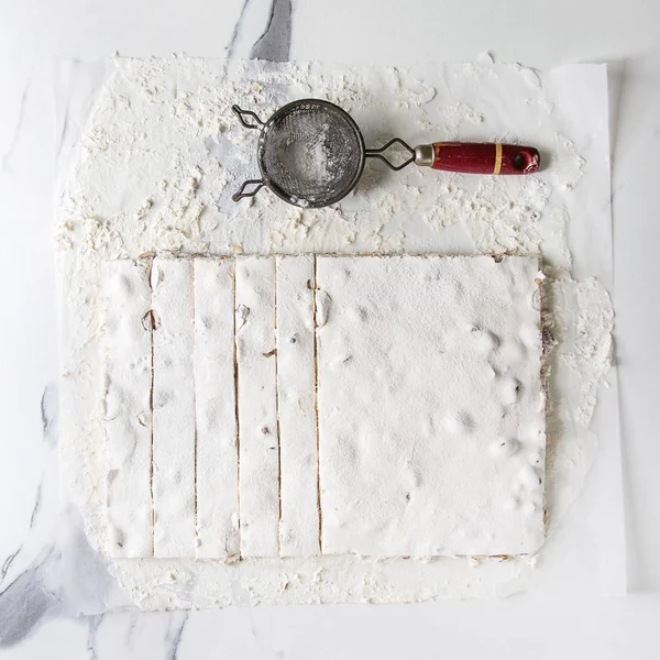 Homemade honey nuts nougat turron sliced on crumpled paper with vintage sieve and sugar powder over white marble background. Flat lay, space. Square image