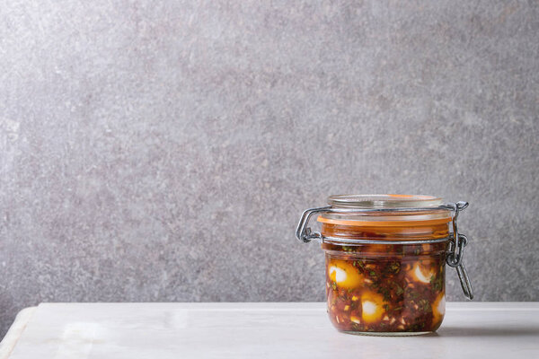 Банка с маринованными перепелиными яйцами в томатном и оливковом соусе с анчоусами и свежей петрушкой на белом мраморном кухонном столе. Копирование пространства
