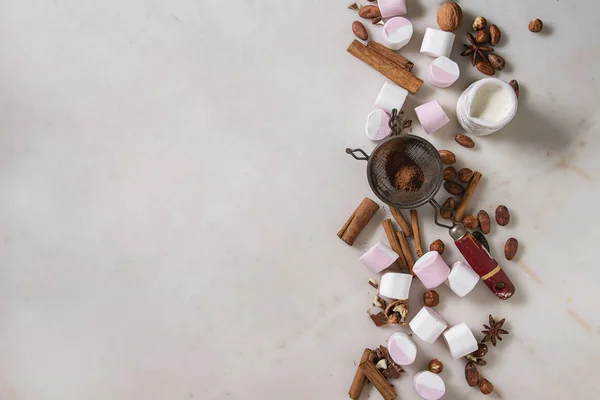 Ingredients for hot chocolate. White pink big marshmallow, cocoa beans, cocoa powder, cinnamon, cream, chopped chocolate, nuts over white marble background. Flat lay, space. Sweet food and drink.