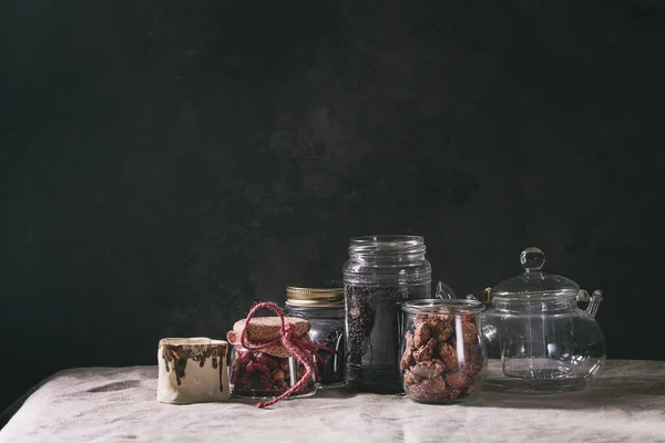 Tea collection. Black dry tea in jar, rose buds, empty glass teapot, cream jug, sweet sugared almond standing on table with linen table cloth.