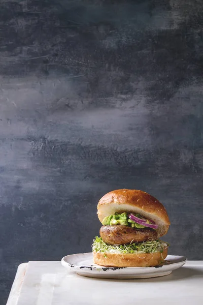Homemade vegan vegetarian burger in wheat bun with portobello mushroom, avocado salsa and sprouts on white ceramic plate over white marble table.