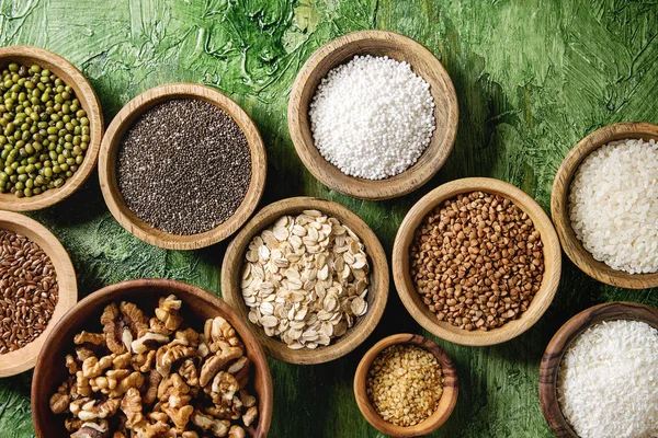 Variety of raw uncooked grains superfood cereal chia linen, sesame, mung bean, walnuts, tapioca, wheat, buckwheat, oatmeal, coconut, rice in wooden bowls over green texture background. Flat lay, space