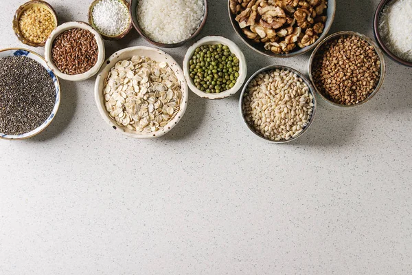 Variety of raw uncooked grains superfood cereal chia, linen, sesame, mung bean, walnuts, tapioca, wheat, buckwheat, oatmeal, coconut, rice in ceramic bowls grey spotted background. Flat lay space — Stock Image Variety Raw Uncooked Grains Superfood Cereal Chia Linen Sesame Mung Stock Picture