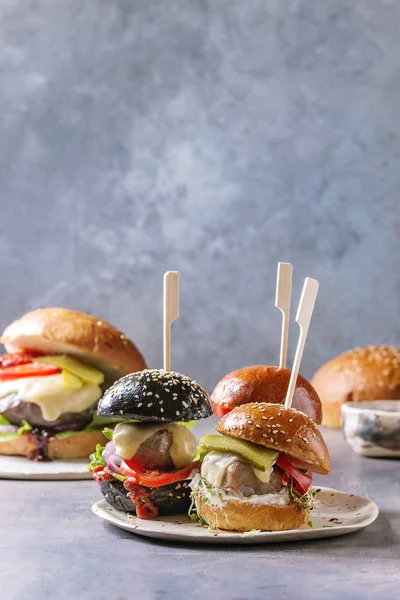 stock image Variety of homemade classic and mini burgers in wheat and black buns with beef and veal cutlets, melted cheese and vegetables on white ceramic board over grey blue table.