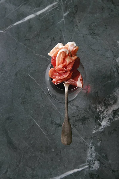 Antipasto meat assorti of sliced jamon, salami, chorizo sausage on fork under glass of red wine over black marble background. Flat lay, space. Appetizer concept