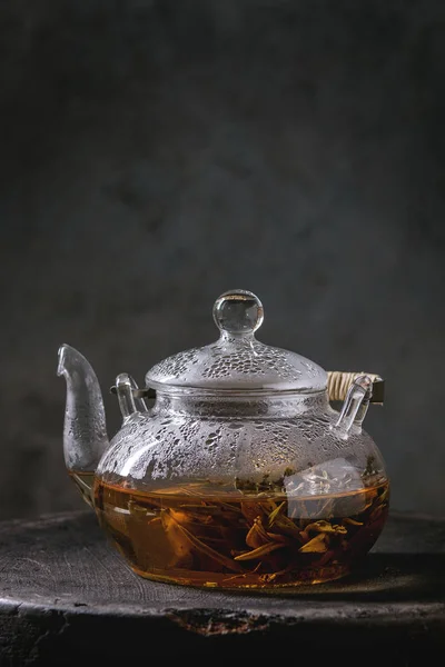 Hot green tea in transparent glass teapot standing on clay tray over black marble table.