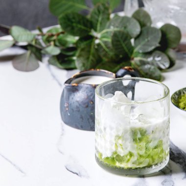 Matcha yeşil latte buzlu çay veya kokteyl cam buz küpleri ile matcha tozu ve süt sürahi beyaz mermer masa, yeşil dallar tarafından dekore edilmiş. Arka plan gri duvara. Kare görüntü