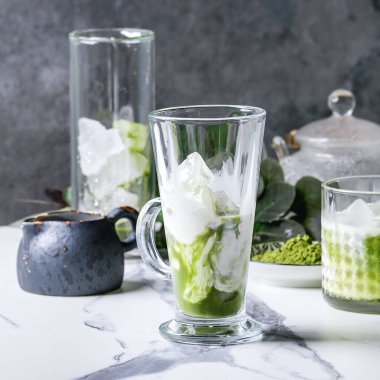 Buzlu latte Matcha yeşil çay veya kokteyl buz küpleri ile üç farklı bardaklarda, matcha toz, sürahi süt beyaz mermer masada yeşil dallar tarafından dekore edilmiş. Arka plan gri duvara. Kare görüntü