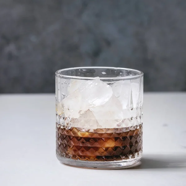 Iced coffee cocktail or frappe with ice cubes in glass on white marble table with grey concrete wall at background. Square image
