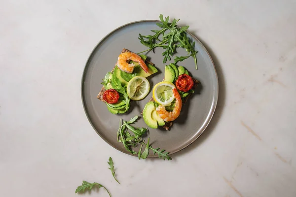Vegetarian sandwich with sliced avocado, sun dried tomatoes, shrimps, arugula served on ceramic plate over white marble background. Flat lay, space