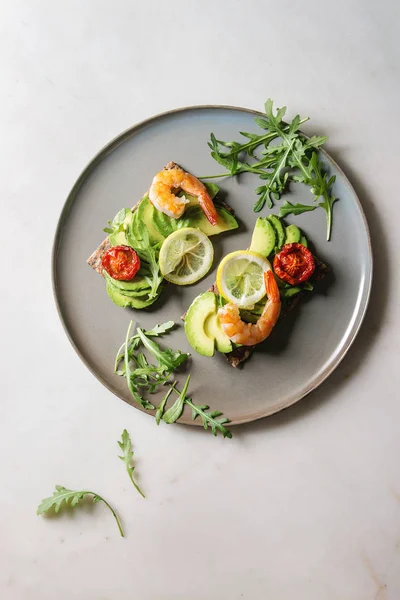 Vegetarian sandwich with sliced avocado, sun dried tomatoes, shrimps, arugula served on ceramic plate over white marble background. Flat lay, space