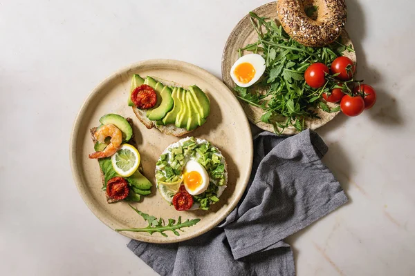Variety of vegetarian sandwiches with sliced avocado, sun dried tomatoes, egg, shrimps, arugula served on ceramic plate over white grey spotted background. Flat lay, space