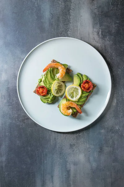 Vegetarian sandwich with sliced avocado, sun dried tomatoes, shrimps, lemon served on ceramic plate over blue texture background. Flat lay, space