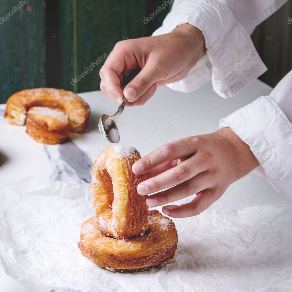 Boy's hand hold homemade puff pastry deep fried donuts or cronuts with sugar standing on crumpled paper over white marble kitchen table. Square image