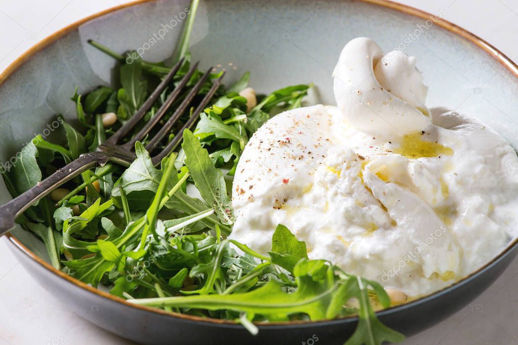 Sliced Italian burrata cheese, fresh arugula salad, pine nuts and olive oil in white ceramic plate on cloth over white marble table. Close up