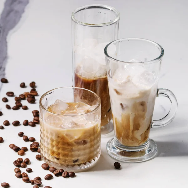 Iced coffee cocktail or frappe with ice cubes and cream served in three different glasses with coffee beans around on white marble table. Square image