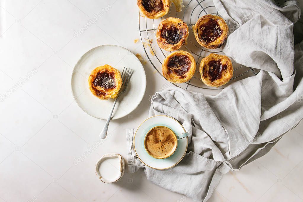 Traditional Portuguese egg tart dessert Pasteis Pastel de nata on cooling rack and ceramic plate with fork, cup of black coffee and jug of cream over white marble background. Flat lay, space